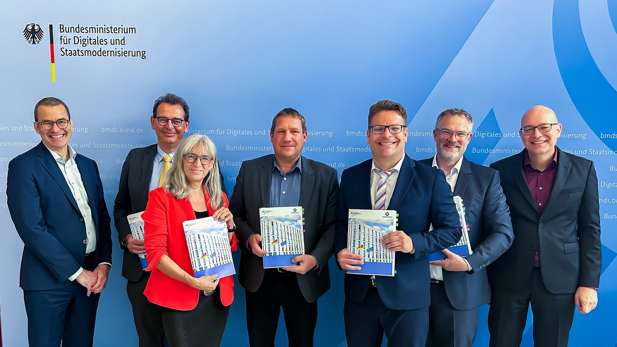 BMDS_Foto Gruppenfoto im Bundesministerium für Digitales und Staatsmodernisierung: Sieben Personen stehen lächelnd vor einer blauen Wand mit Ministeriumslogo. In der Mitte halten mehrere von ihnen eine Broschüre mit dem Foto eines Regierungsgebäudes.
